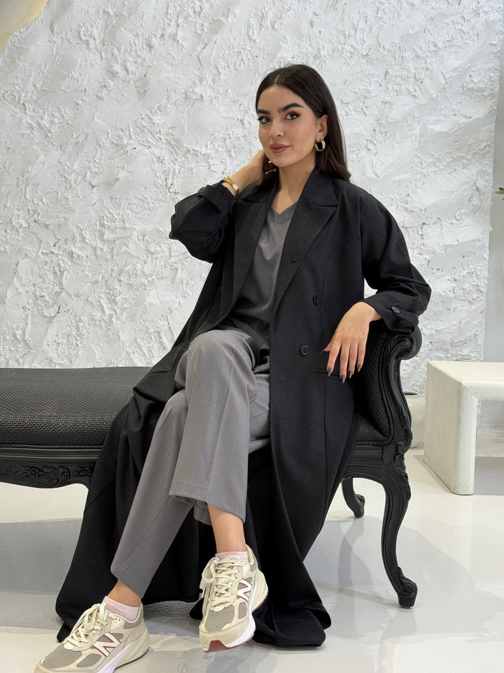 Woman in a Black Jookh Abaya Set sitting on a white bench with a textured white wall background