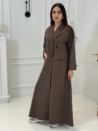 Woman wearing a long cocoa brown Jookh Abaya Set standing in front of a white wall with a gold-framed mirror.