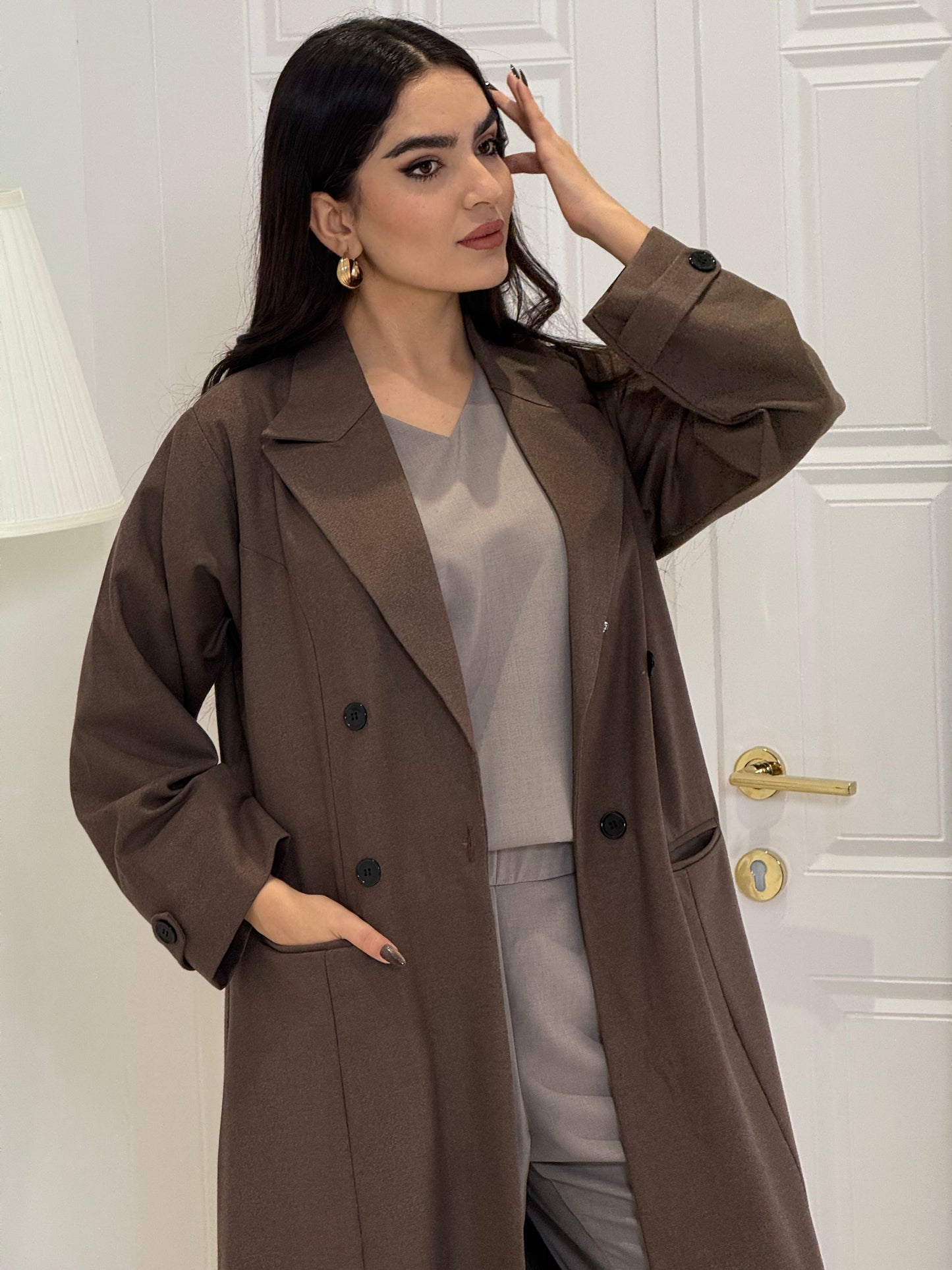 Woman wearing a long cocoa brown Jookh Abaya Set standing in front of a white wall with a gold-framed mirror.