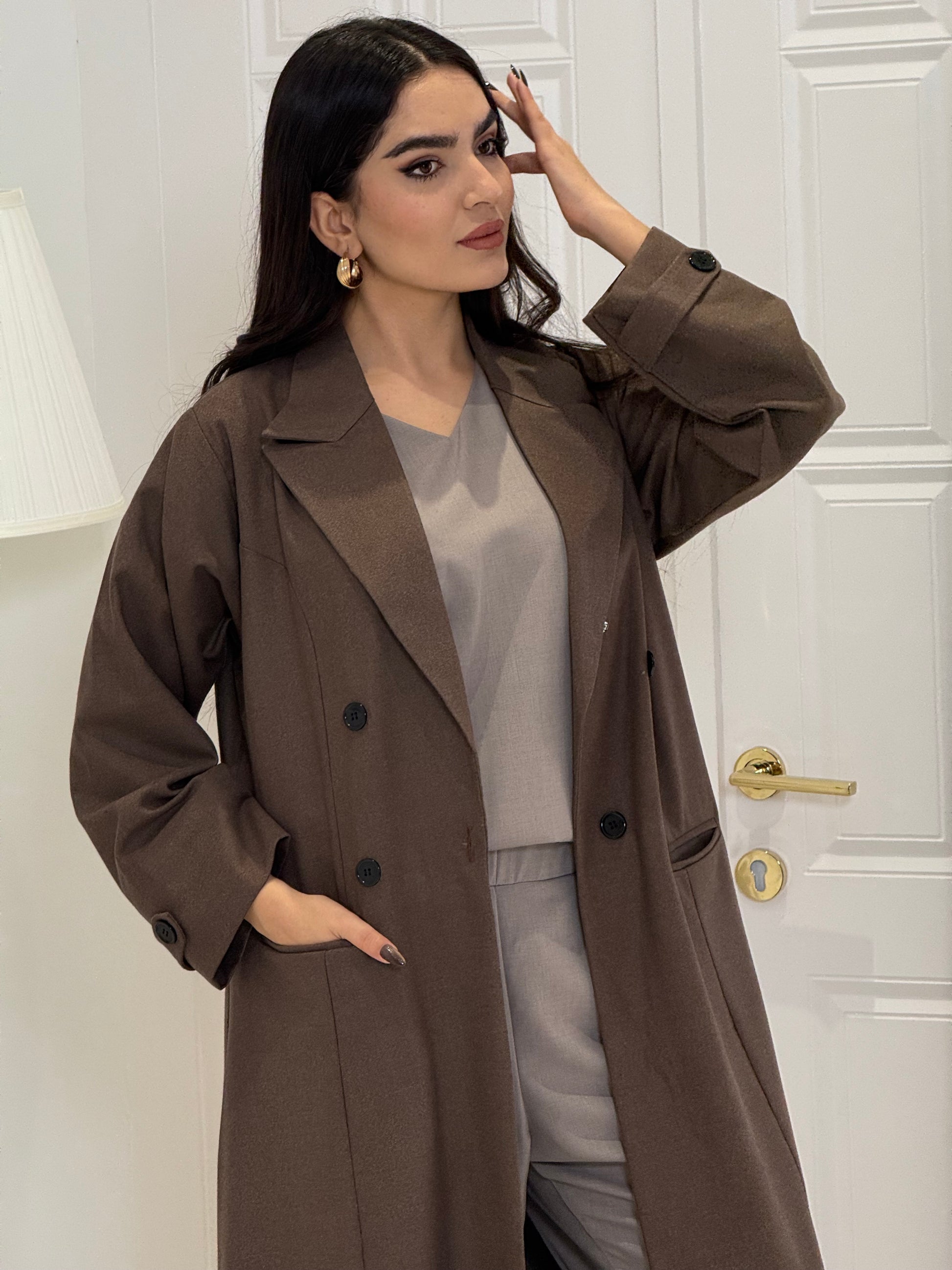 Woman wearing a long cocoa brown Jookh Abaya Set standing in front of a white wall with a gold-framed mirror.