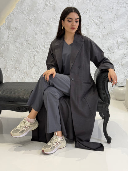 Woman wearing a long dark grey Jookh Abaya Set standing in front of a white wall with a gold-framed mirror.