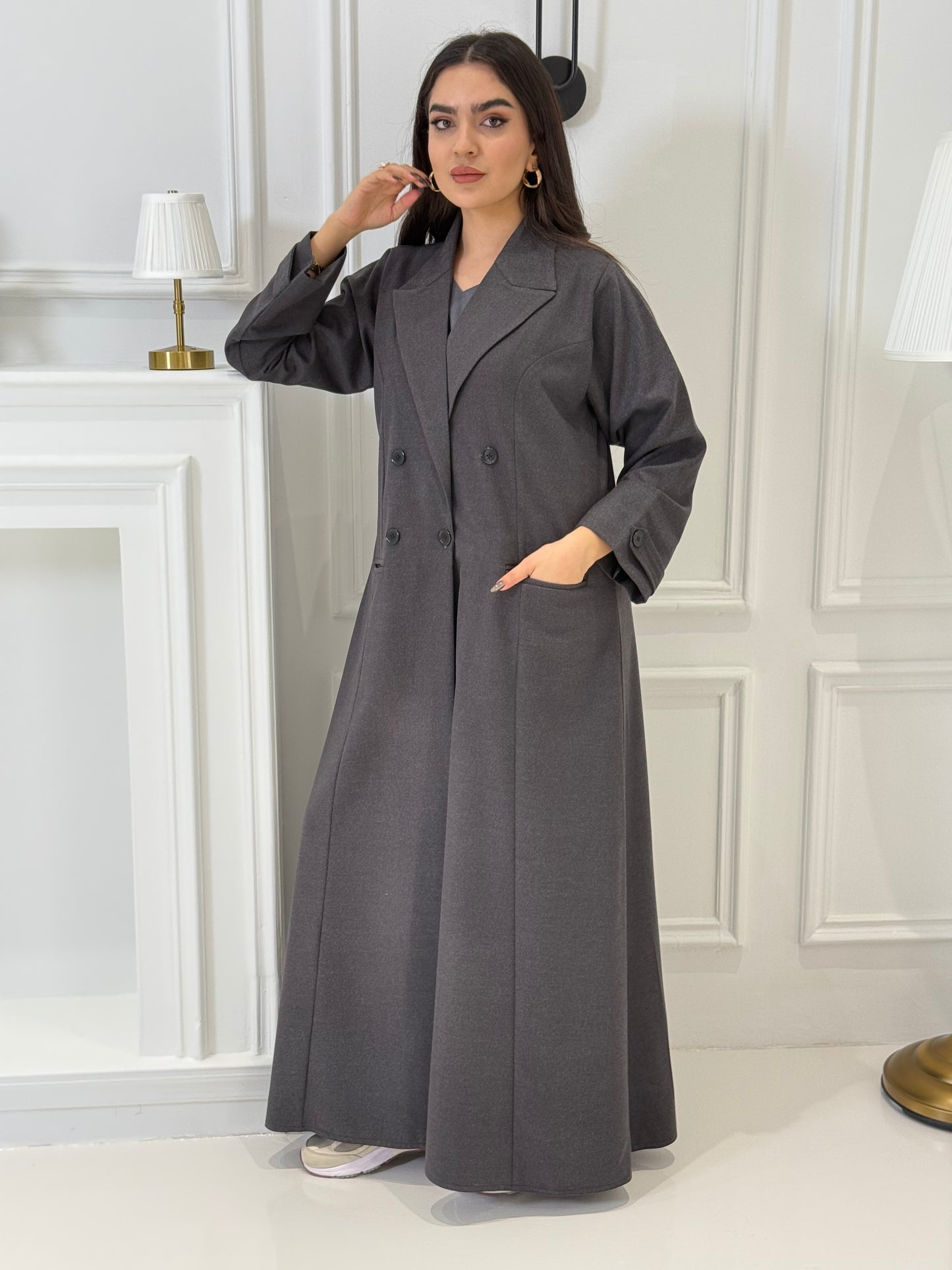 Woman wearing a long dark grey Jookh Abaya Set standing in front of a white wall with a gold-framed mirror.