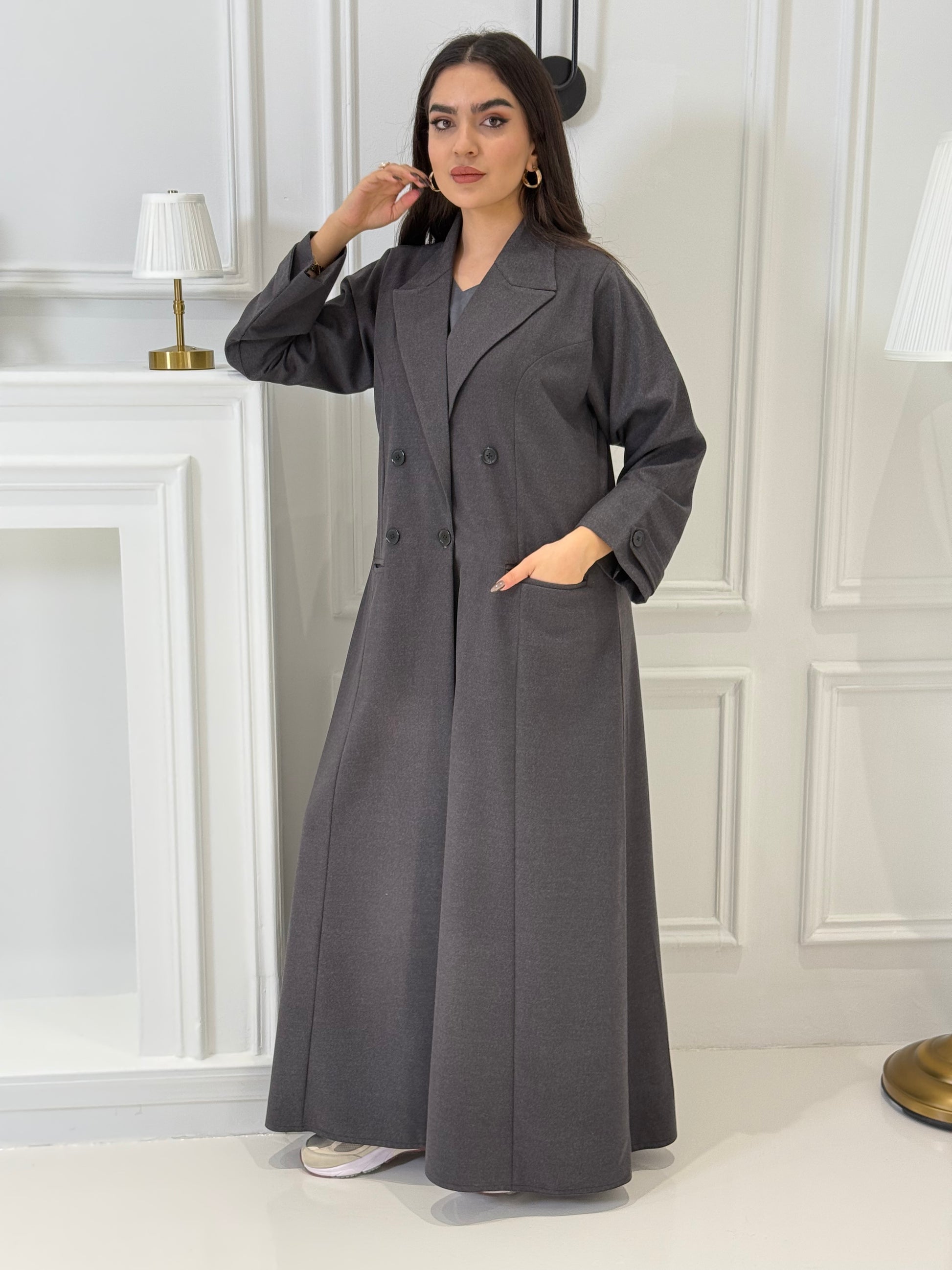 Woman wearing a long dark grey Jookh Abaya Set standing in front of a white wall with a gold-framed mirror.