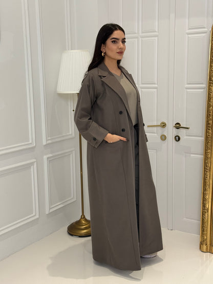 Woman wearing a long dark khaki Jookh Abaya Set standing in front of a white wall with a gold-framed mirror.