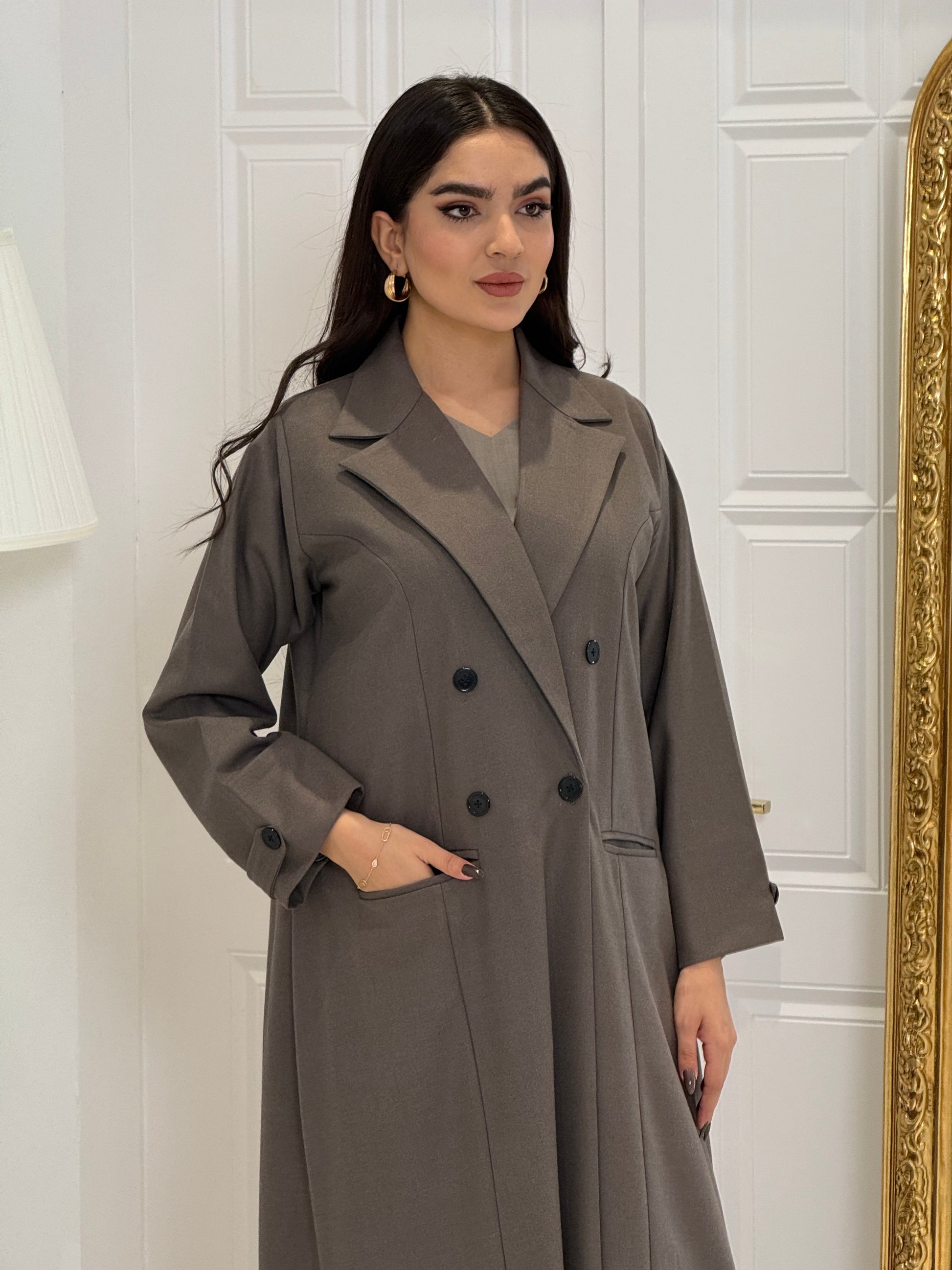 Woman wearing a long dark khaki Jookh Abaya Set standing in front of a white wall with a gold-framed mirror.