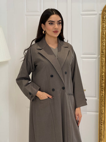 Woman wearing a long dark khaki Jookh Abaya Set standing in front of a white wall with a gold-framed mirror.