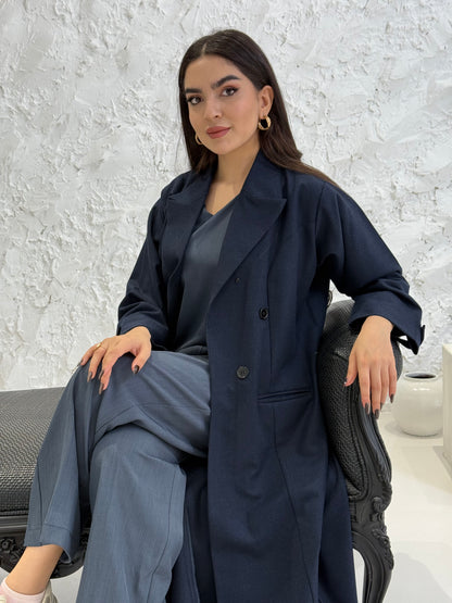 Woman wearing a long navy blue Jookh Abaya Set standing in front of a white wall with a gold-framed mirror.