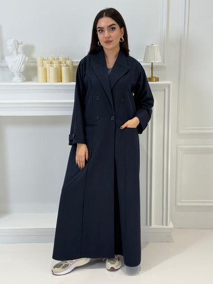 Woman wearing a long navy blue Jookh Abaya Set standing in front of a white wall with a gold-framed mirror.