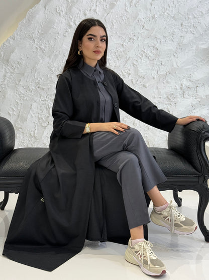 Woman sitting on a chair wearing a black coat jookh abaya and gray pants, holding a black handbag.