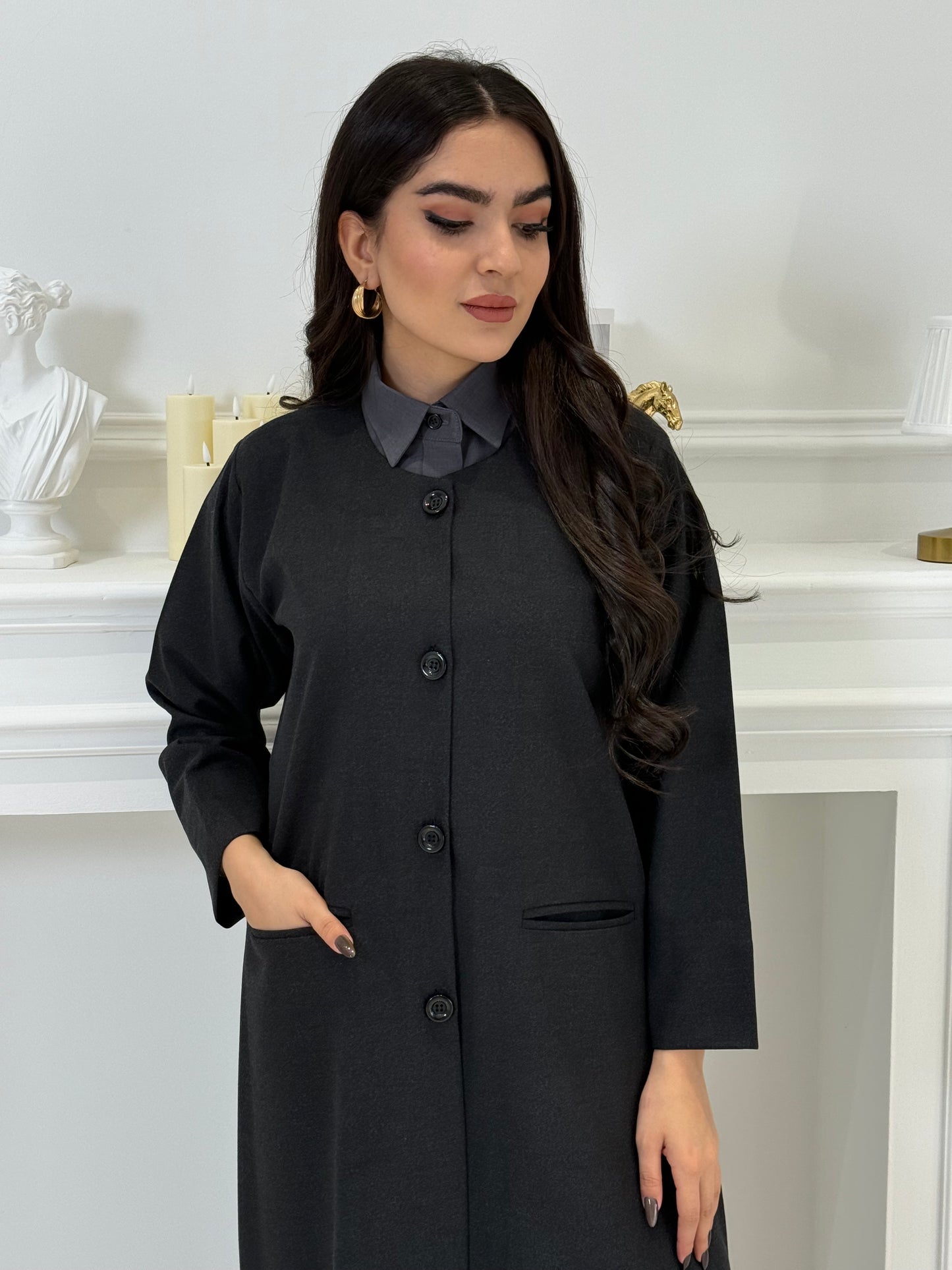 Woman wearing a black coat jookh abaya and gray pants, holding a black handbag.