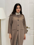 Woman wearing a long light brown jookh  coat abaya in a room with white walls and doors.