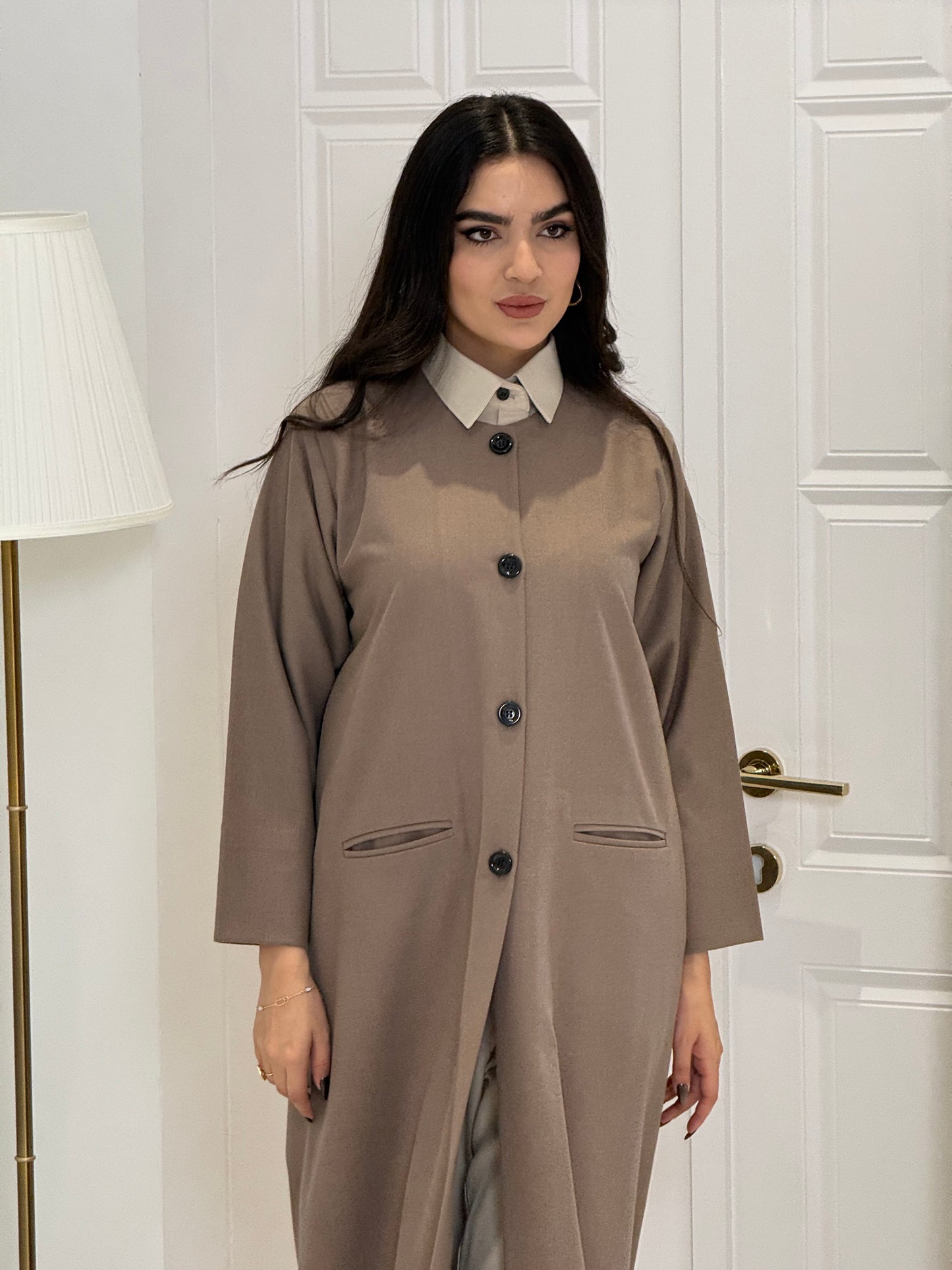 Woman wearing a long light brown jookh  coat abaya in a room with white walls and doors.