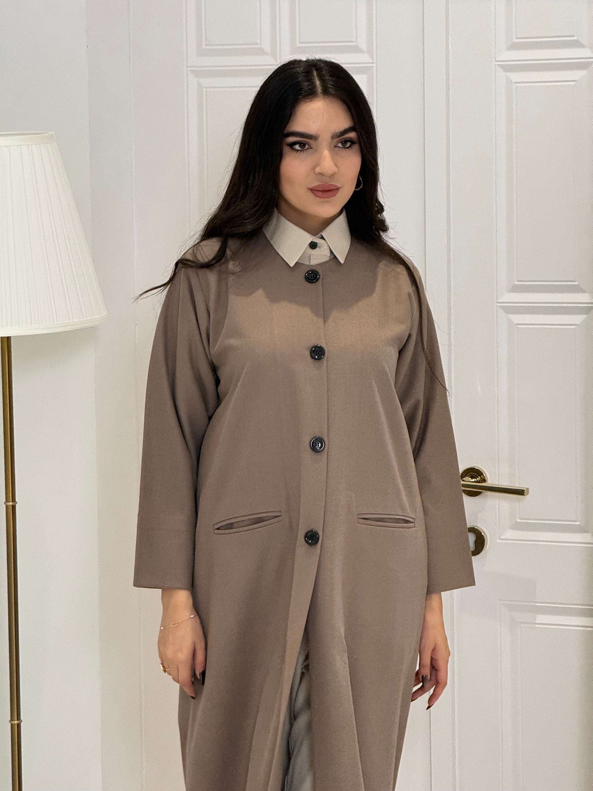 Woman wearing a long light brown jookh  coat abaya in a room with white walls and doors.