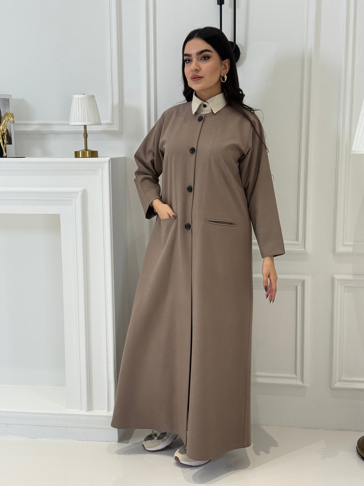 Woman wearing a long light brown jookh  coat abaya in a room with white walls and doors.