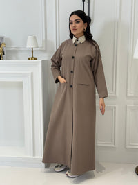 Woman wearing a long light brown jookh  coat abaya in a room with white walls and doors.