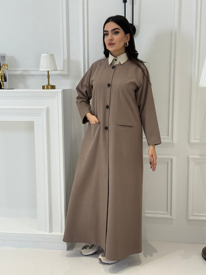 Woman wearing a long light brown jookh  coat abaya in a room with white walls and doors.
