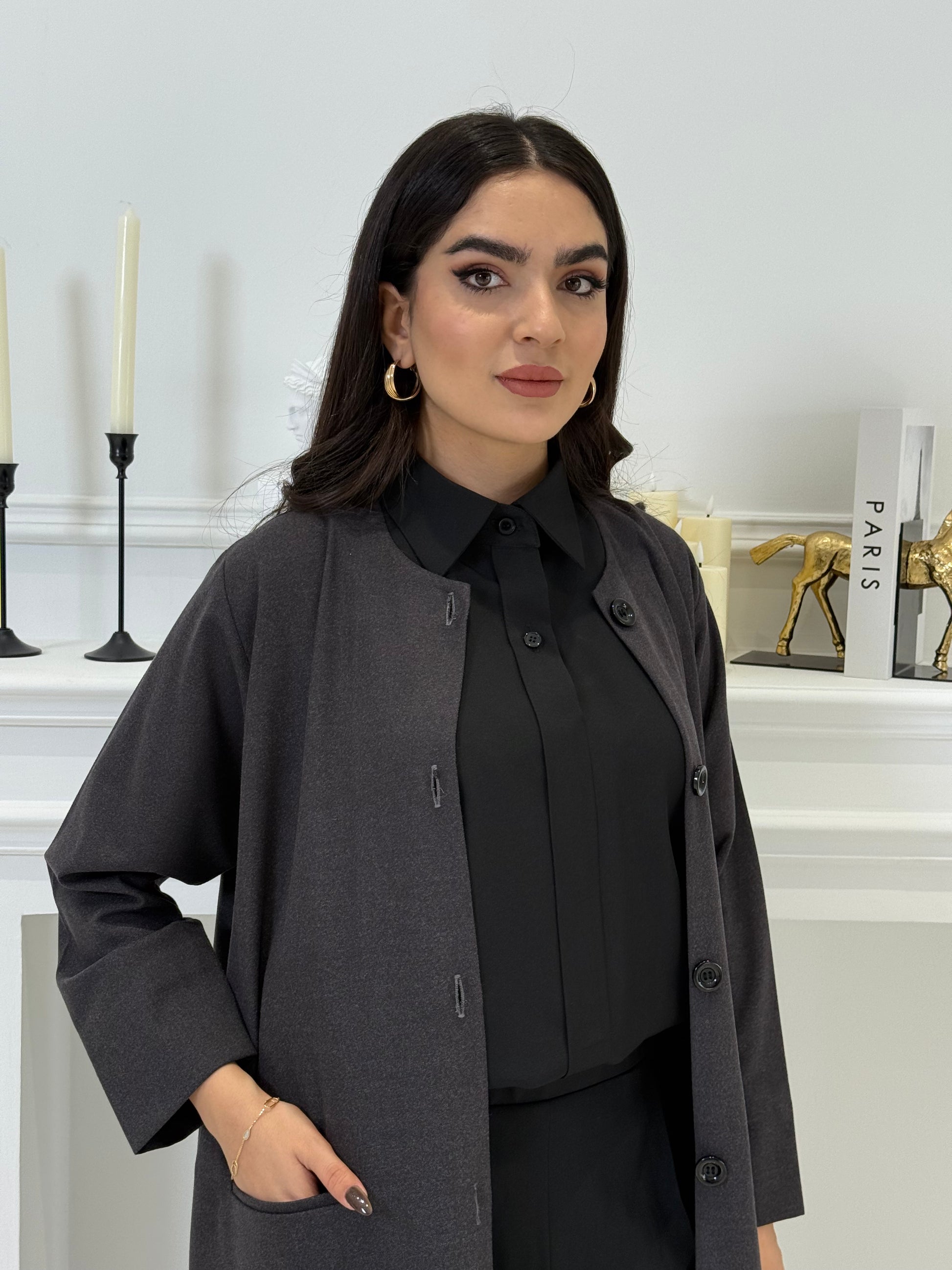 Woman wearing a dark grey coat jookh abaya and gray pants, holding a black handbag.