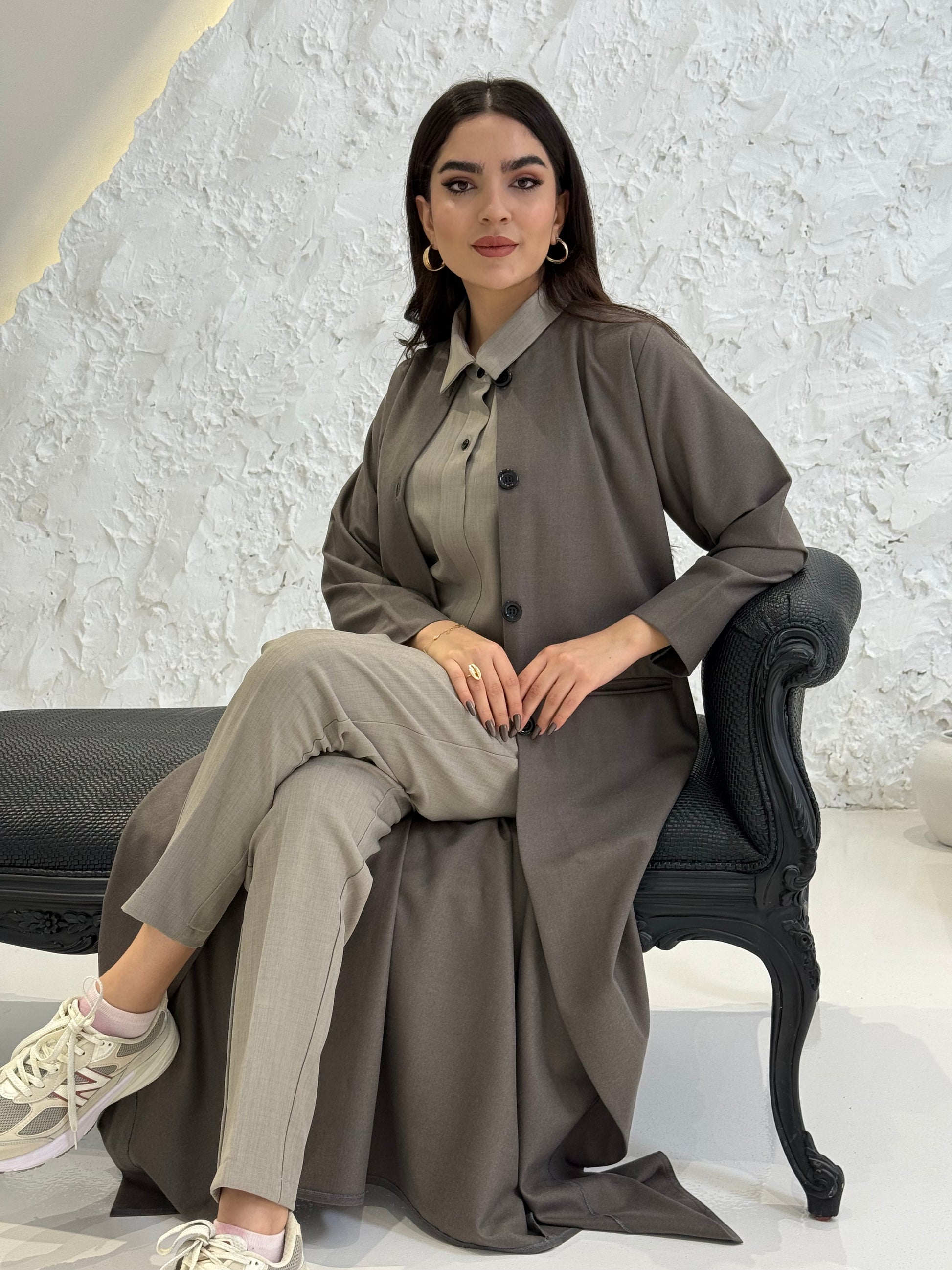 Woman wearing a dark taupe coat jookh abaya and gray pants, holding a black handbag.