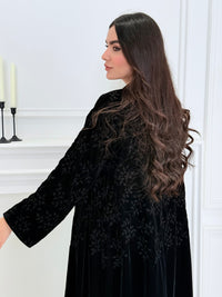 A woman wearing a long Black Velvet Embroidered Abaya standing against a white decoration 