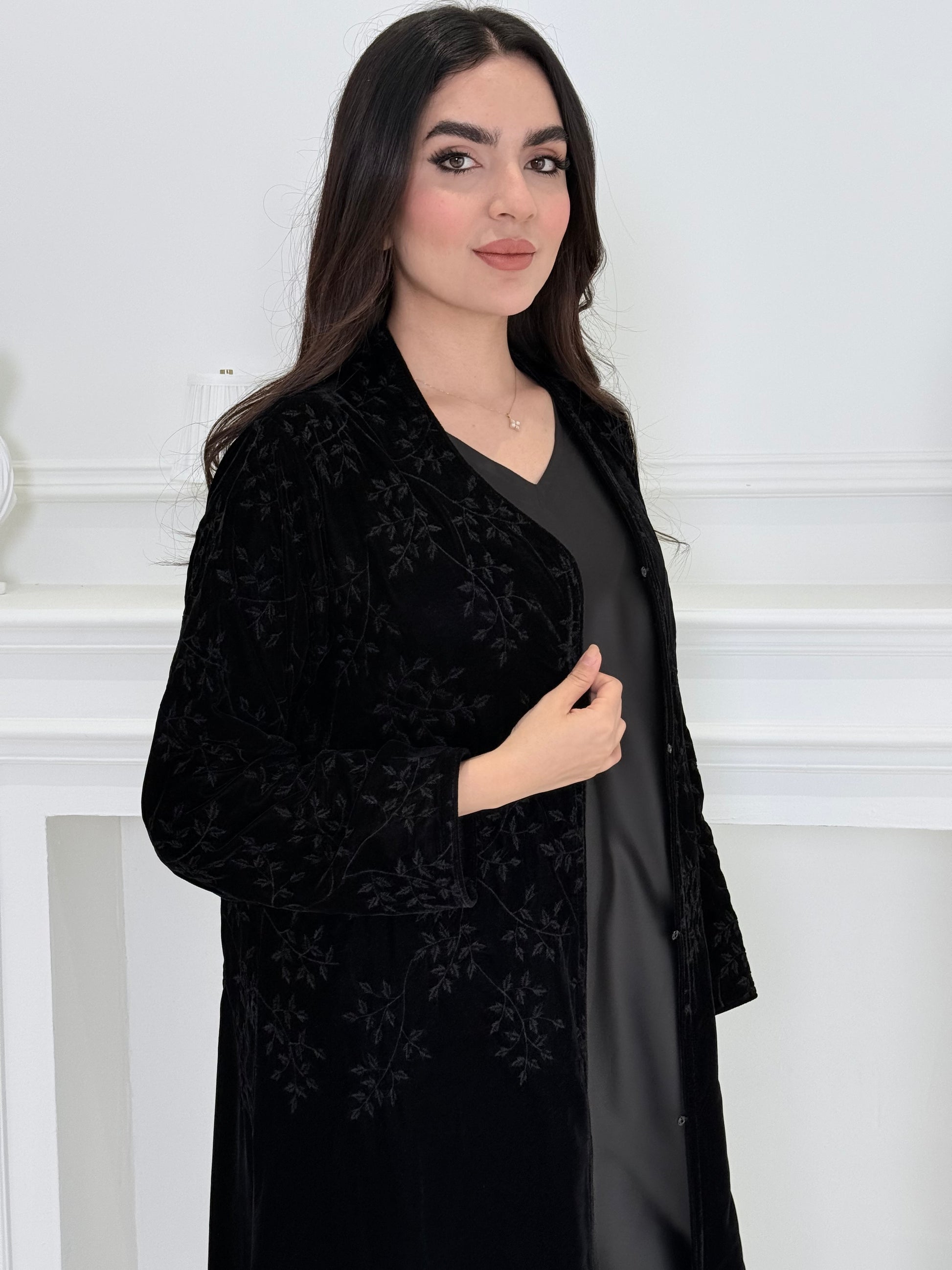 A woman wearing a long Black Velvet Embroidered Abaya standing against a white decoration 