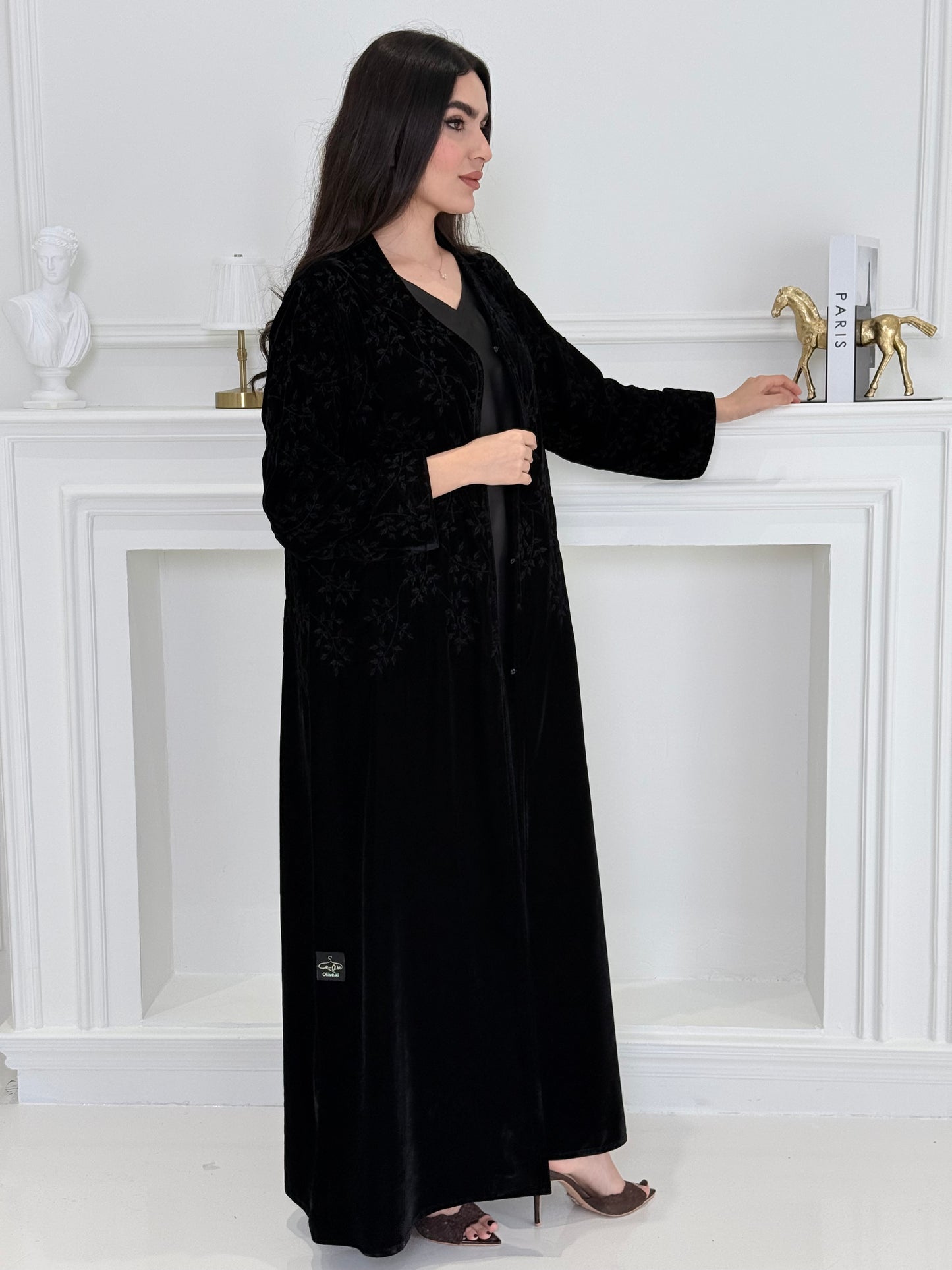 A woman wearing a long Black Velvet Embroidered Abaya standing against a white decoration 