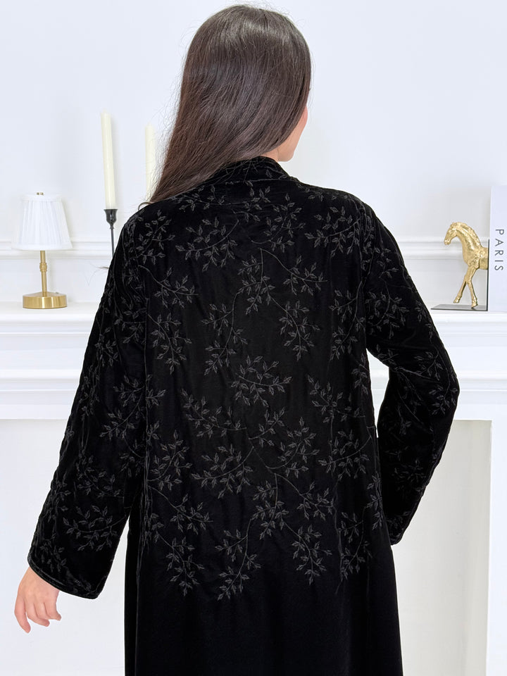 A woman wearing a long Black Velvet Embroidered Abaya standing against a white decoration 