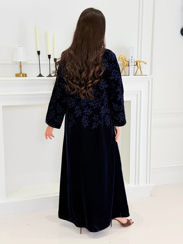 A woman wearing a long navy blue Velvet Embroidered Abaya standing against a white decoration 