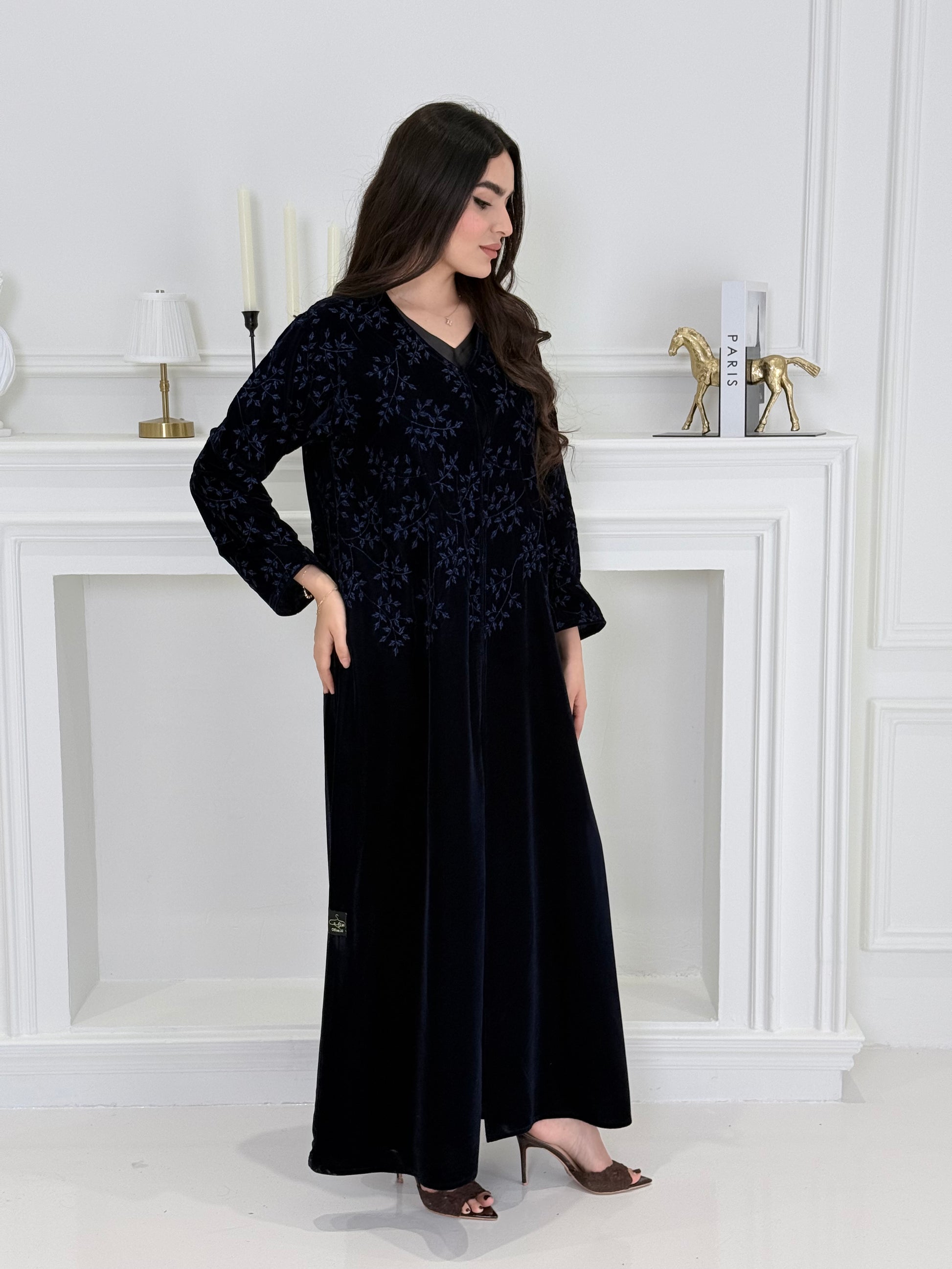 A woman wearing a long navy blue Velvet Embroidered Abaya standing against a white decoration 