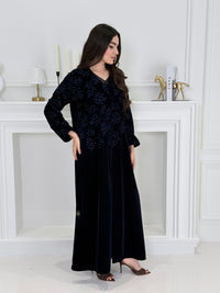 A woman wearing a long navy blue Velvet Embroidered Abaya standing against a white decoration 