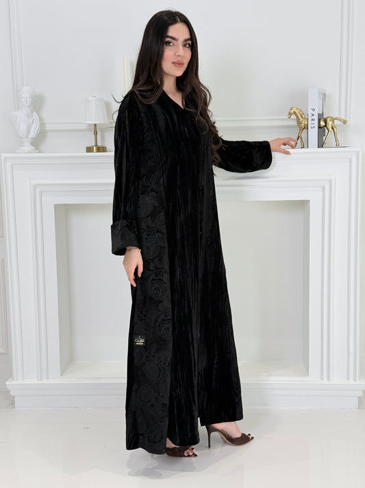 woman wearing a dark black plissé velvet abaya coat standing in front of a white door with gold handles.