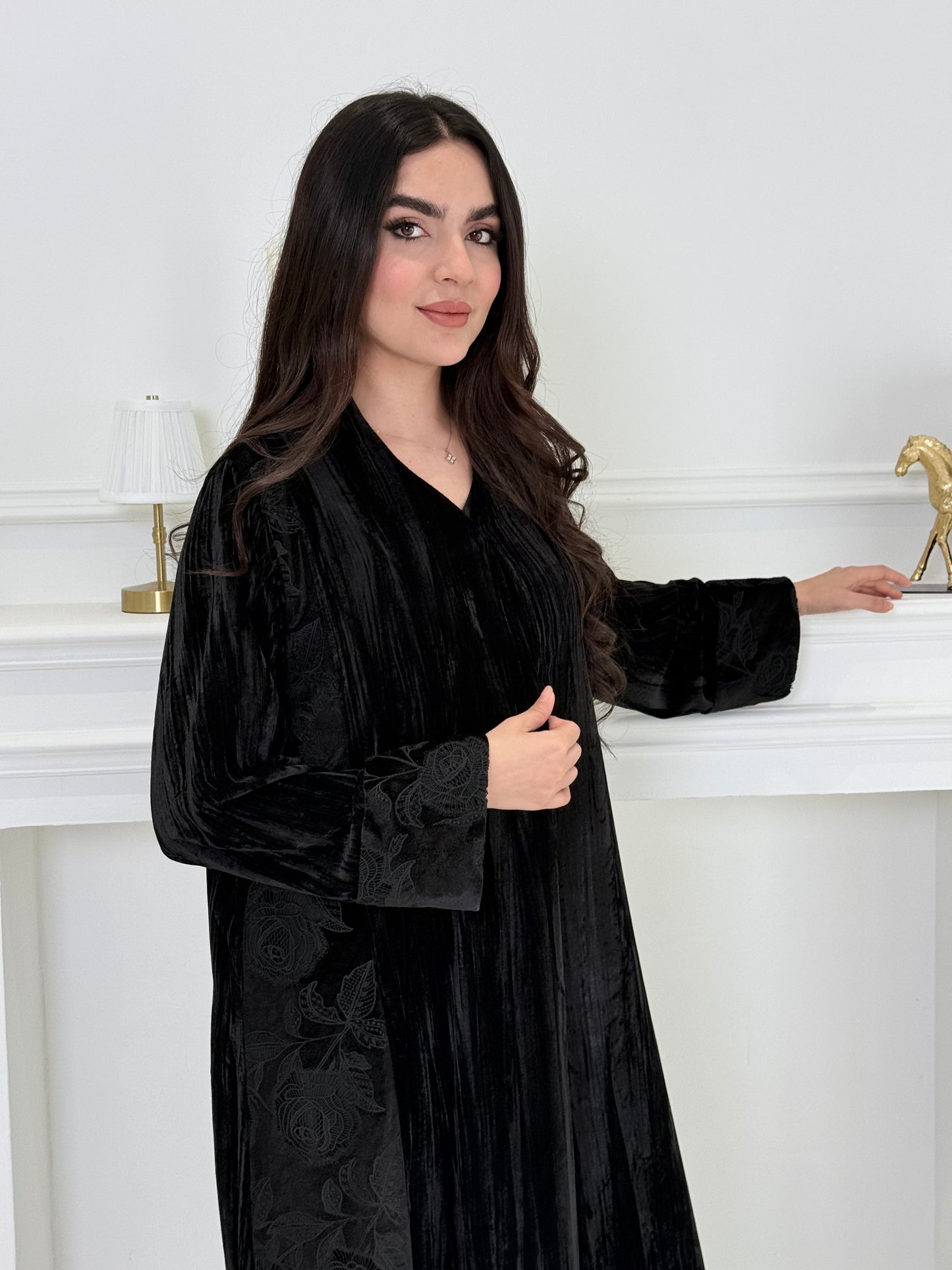 woman wearing a dark black plissé velvet abaya coat standing in front of a white door with gold handles.