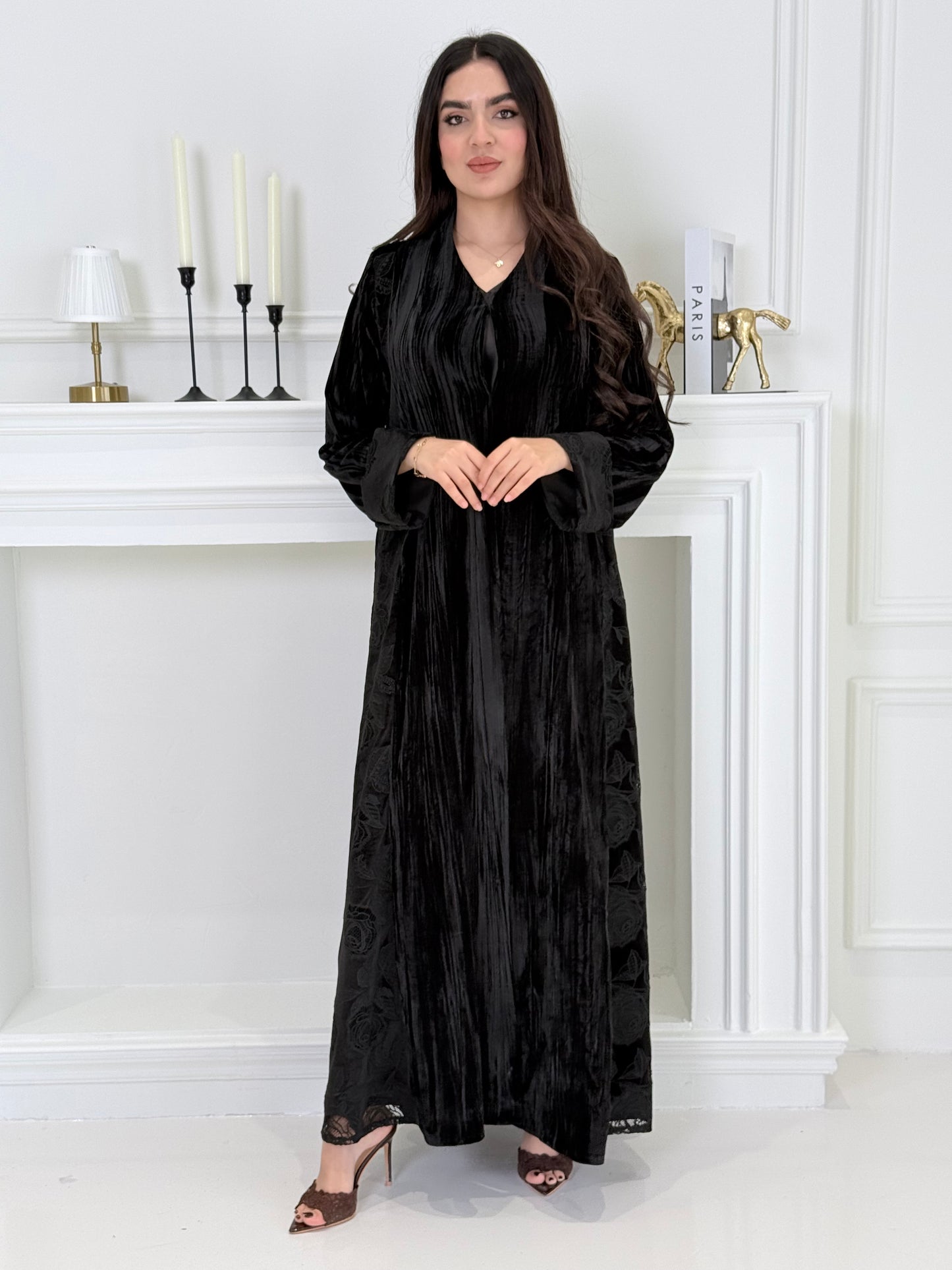 woman wearing a dark black plissé velvet abaya coat standing in front of a white door with gold handles.