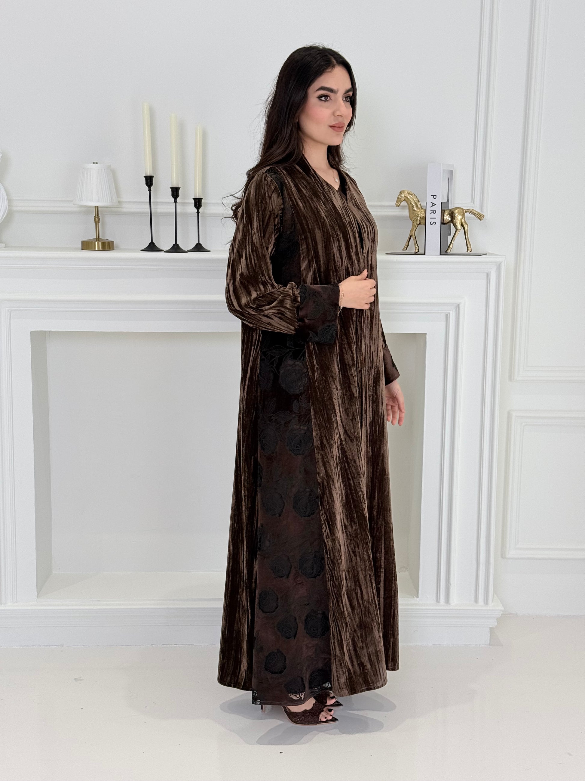 woman wearing a dark brown plissé velvet abaya coat standing in front of a white door with gold handles.