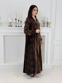 woman wearing a dark brown plissé velvet abaya coat standing in front of a white door with gold handles.