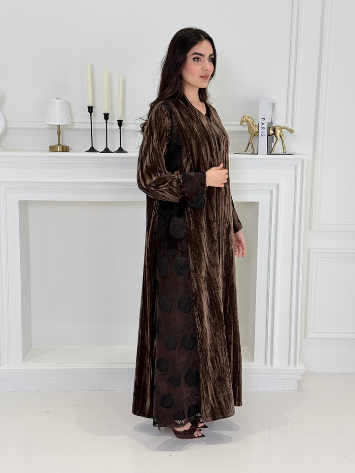 woman wearing a dark brown plissé velvet abaya coat standing in front of a white door with gold handles.
