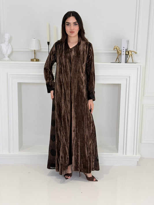 woman wearing a dark brown plissé velvet abaya coat standing in front of a white door with gold handles.