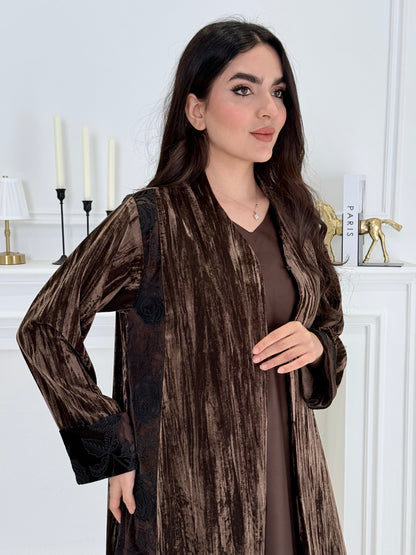 woman wearing a dark brown plissé velvet abaya coat standing in front of a white door with gold handles.