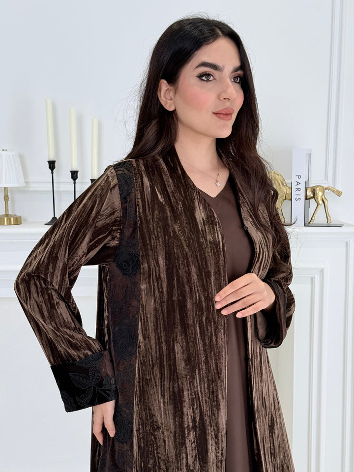woman wearing a dark brown plissé velvet abaya coat standing in front of a white door with gold handles.