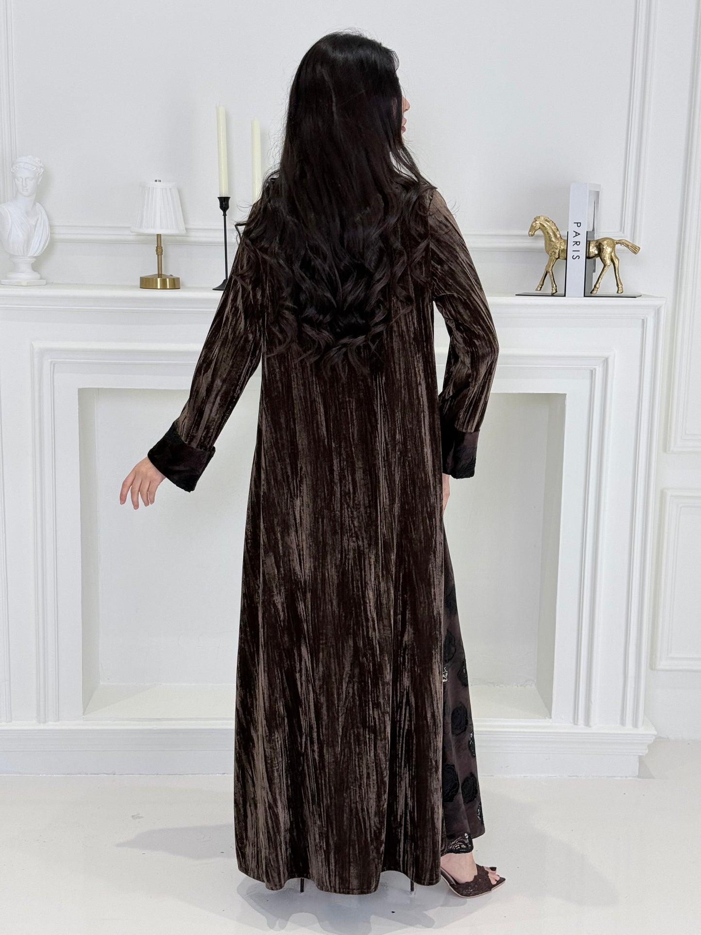 woman wearing a dark brown plissé velvet abaya coat standing in front of a white door with gold handles.