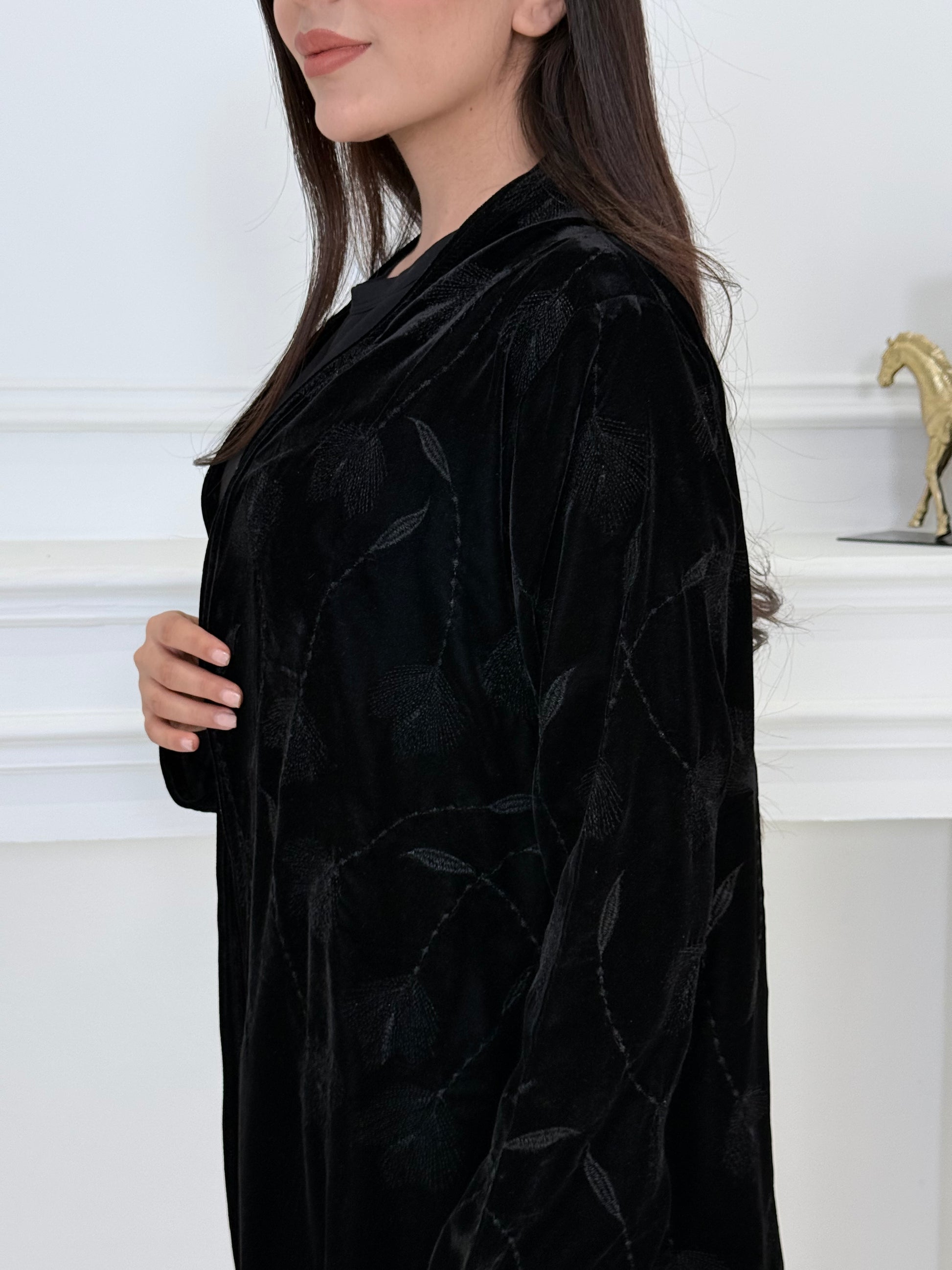 Woman wearing a premium luxury black Floral Velvet Abaya dress with floral embellishments in a white room.