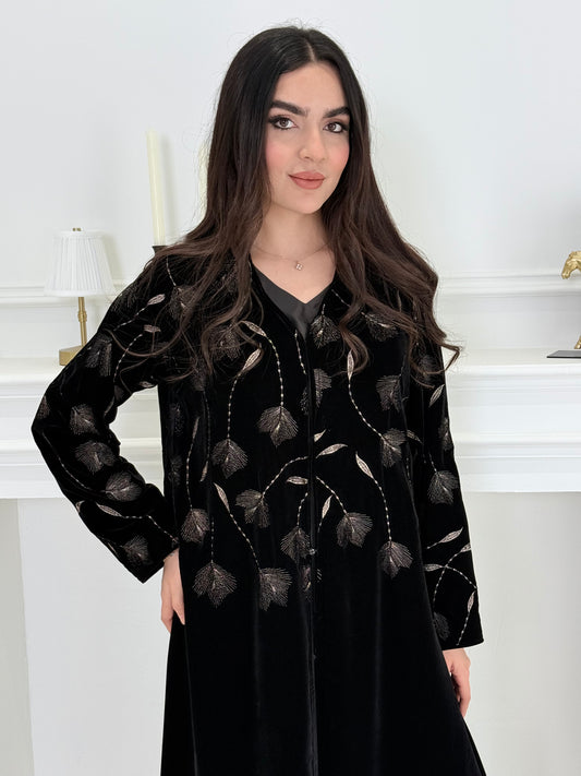 Woman wearing a premium luxury black and Gold Floral Velvet Abaya dress with floral embellishments in a white room.