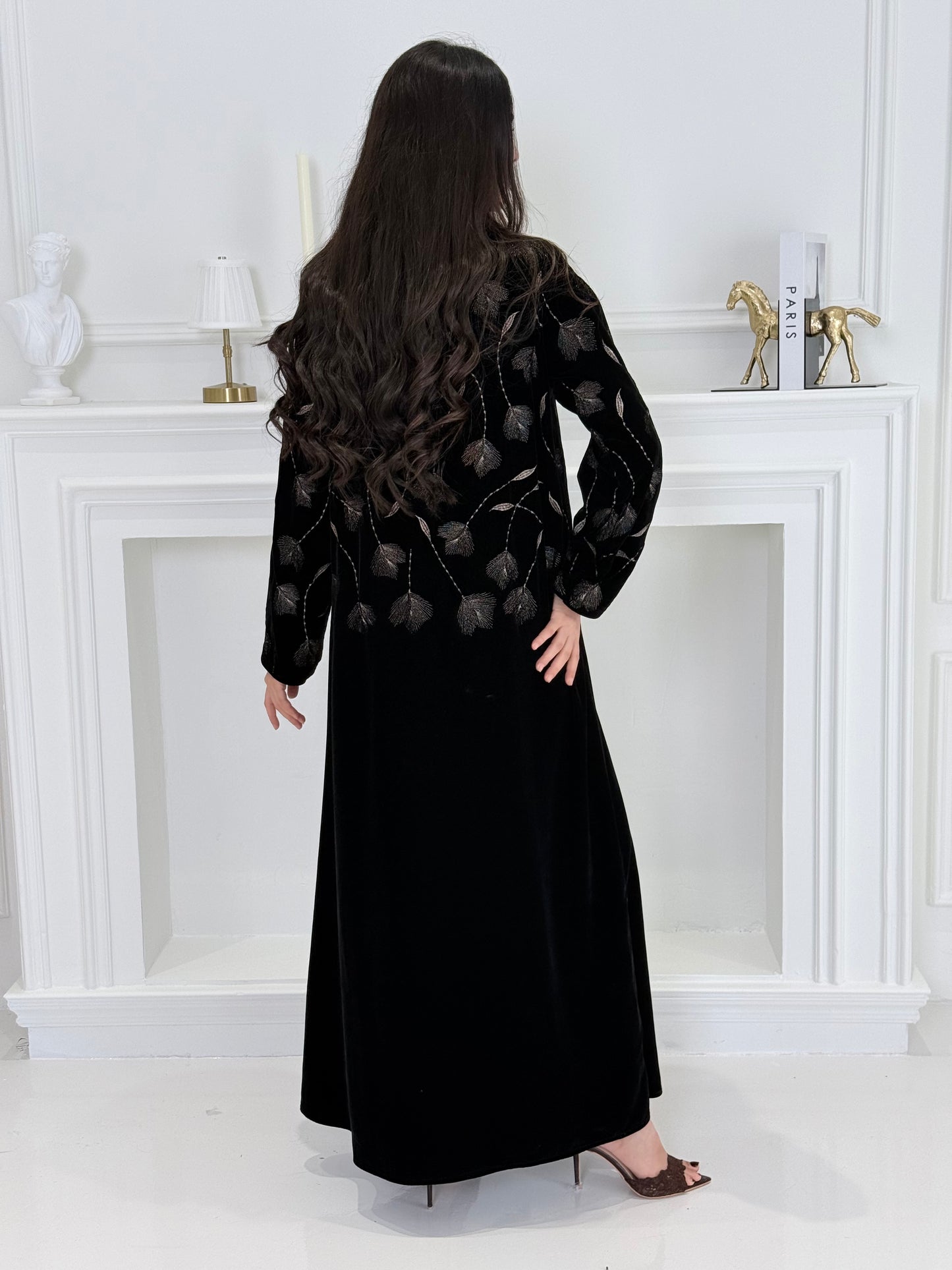 Woman wearing a premium luxury black and Gold Floral Velvet Abaya dress with floral embellishments in a white room.