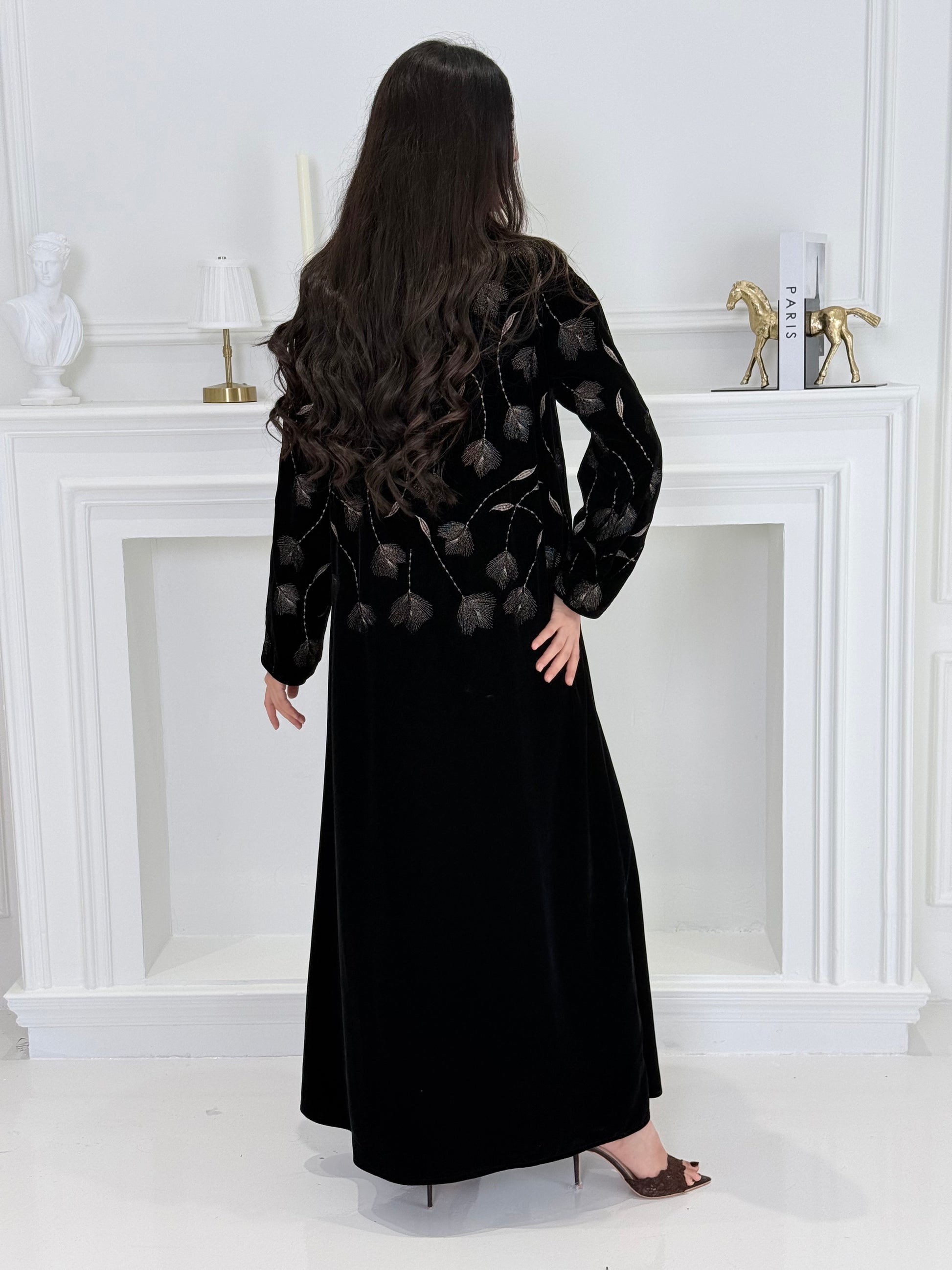 Woman wearing a premium luxury black and Gold Floral Velvet Abaya dress with floral embellishments in a white room.