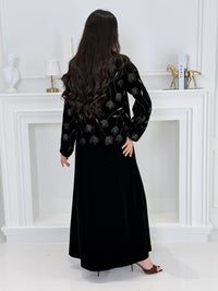 Woman wearing a premium luxury black and Gold Floral Velvet Abaya dress with floral embellishments in a white room.