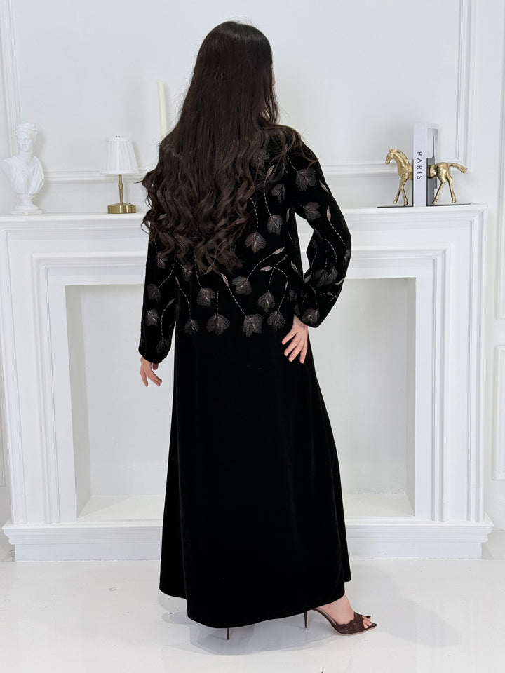 Woman wearing a premium luxury black and Gold Floral Velvet Abaya dress with floral embellishments in a white room.