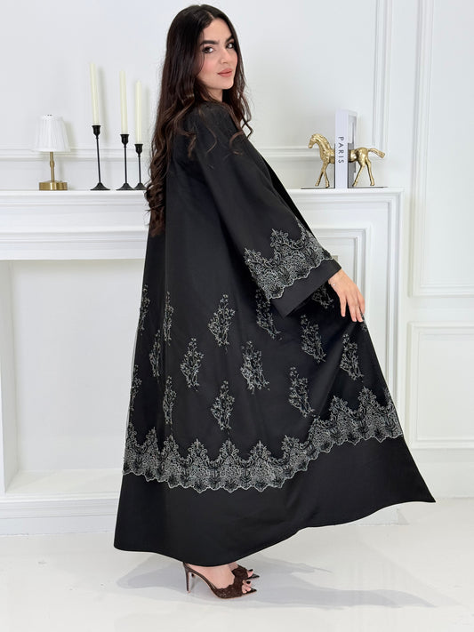 Woman wearing a Premium Silver Embroidered Black Abaya dress with lace details made from Nida fabric and matchhing sheila in a white room.