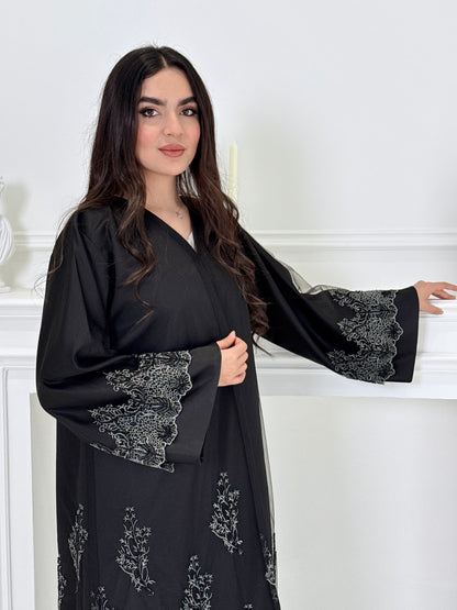 Woman wearing a Premium Silver Embroidered Black Abaya dress with lace details made from Nida fabric and matchhing sheila in a white room.