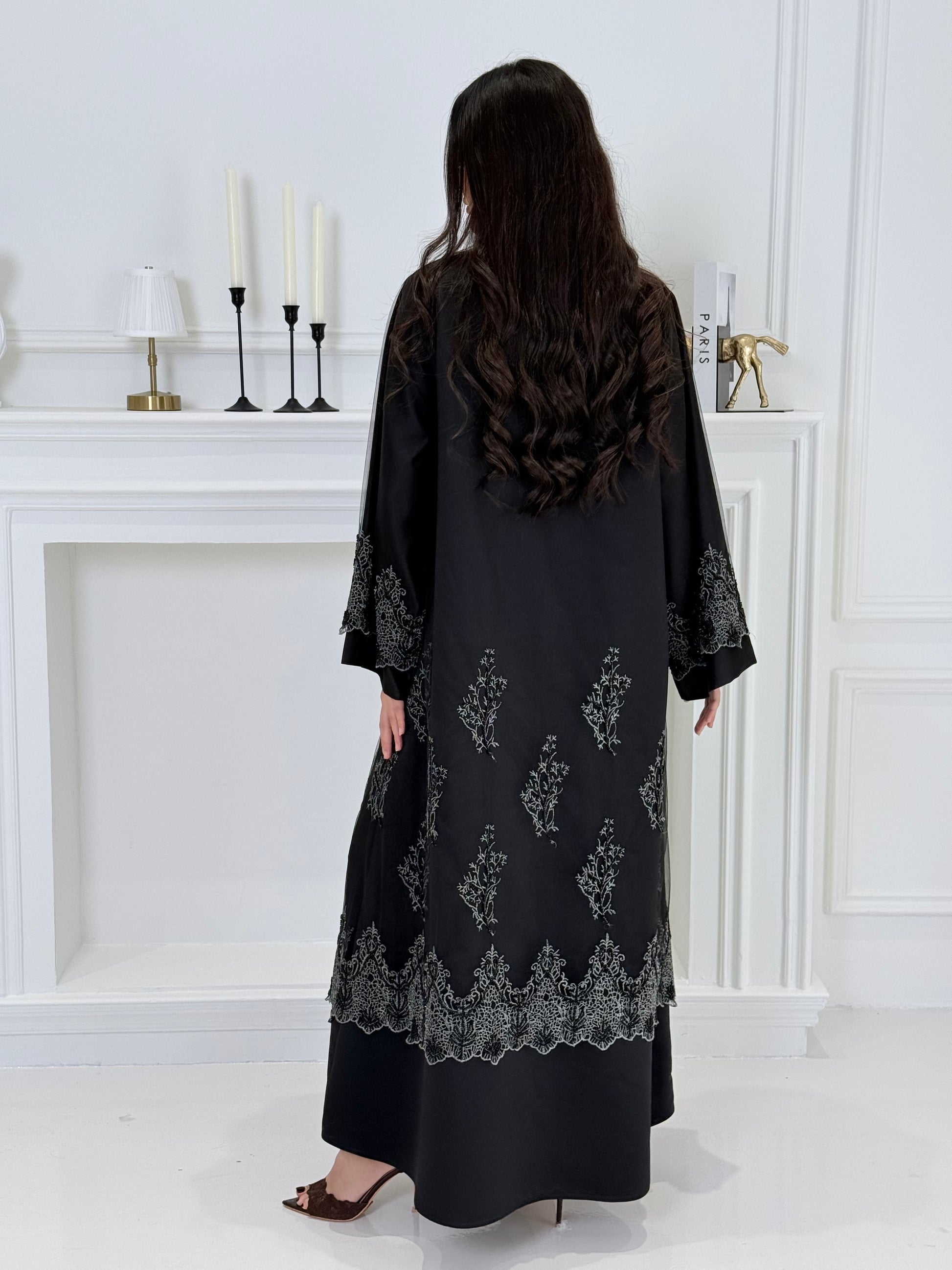 Woman wearing a Premium Silver Embroidered Black Abaya dress with lace details made from Nida fabric and matchhing sheila in a white room.
