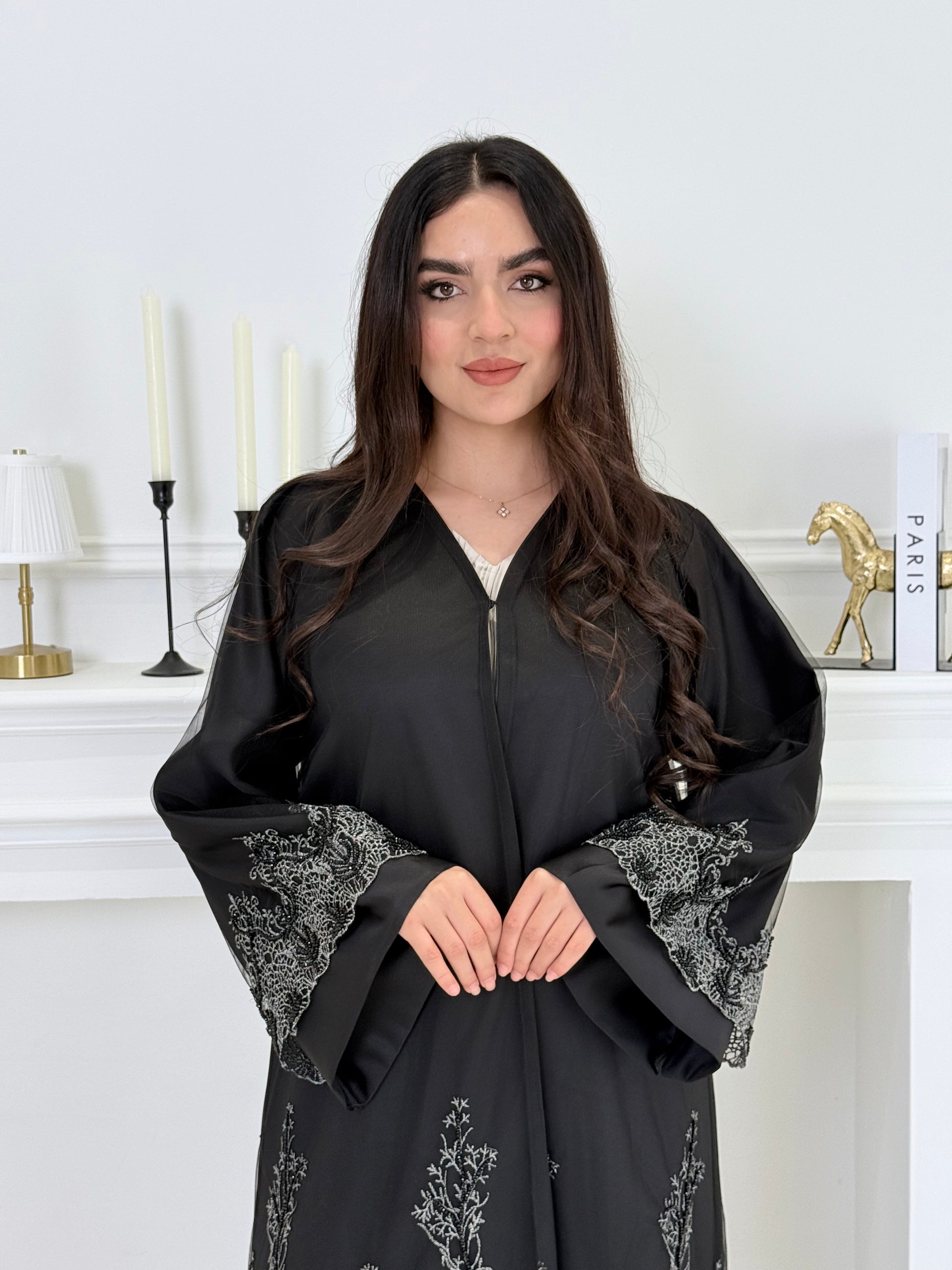 Woman wearing a Premium Silver Embroidered Black Abaya dress with lace details made from Nida fabric and matchhing sheila in a white room.