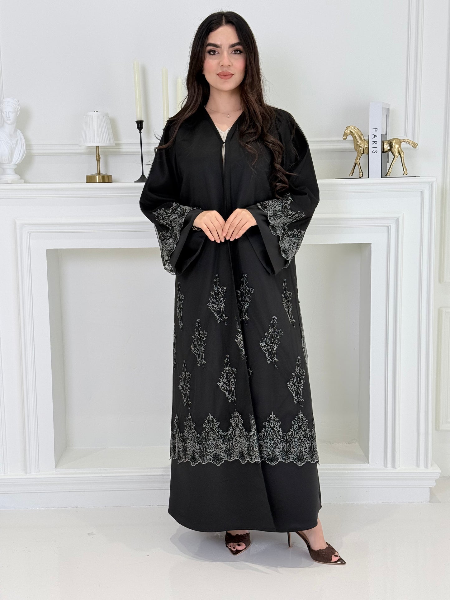 Woman wearing a Premium Silver Embroidered Black Abaya dress with lace details made from Nida fabric and matchhing sheila in a white room.